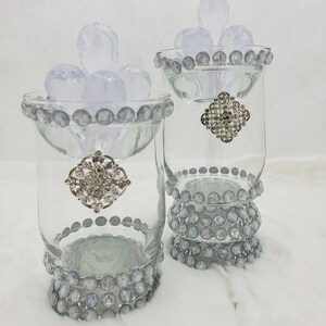Puede incluir: Dos candelabros de vidrio transparente con adornos plateados y pedrería iridiscente alrededor del borde y la base. Los candelabros están llenos de candelabros de vidrio transparente en forma de lágrima.