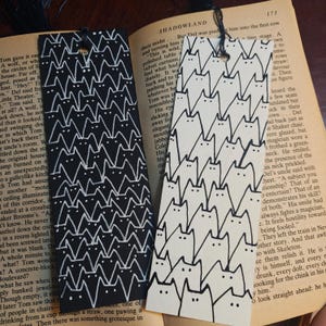 Puede incluir: Dos marcapáginas rectangulares con un patrón repetitivo de gatos. Uno es negro con gatos dibujados en blanco y el otro es color crema con gatos dibujados en negro. Cada marcapáginas tiene un agujero en la parte superior con una borla.
