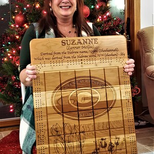 May include: A wooden Green Bay Packers cribbage board with a personalized inscription that reads "SUZANNE (Zanner McGee) Derived from the Hebrew name שושנה (Shoshannah). Which was derived from the Hebrew word, שושן (shoshan) - lily". The board features the Green Bay Packers logo and the words "Green Bay Packers" and "Est. 1919".