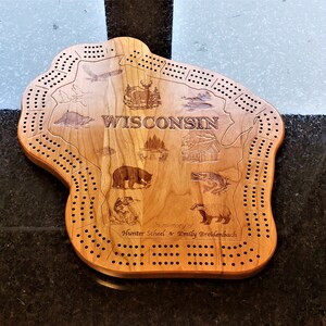 May include: A wooden cribbage board shaped like the state of Wisconsin. The board is engraved with the word "Wisconsin" and various images of wildlife, including a bear, a fish, and a deer. The board also includes the text "In memory of Hunter Scheel & Emily Breidenbach".