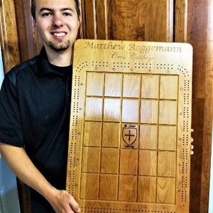 May include: A personalized wooden cribbage board with the name "Matthew Roggemann" and the text "Cribbage" engraved on it. The board has a traditional design with a grid of squares and a scoring track around the perimeter. A small shield with a cross is engraved in the center of the board.