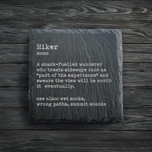 May include: A black slate coaster with the definition of "Hiker" in white text. The definition describes a snack-fuelled wanderer who embraces rain and believes the view is worth it. The coaster is on a dark wood surface.