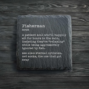 Puede incluir: Un posavasos de pizarra gris oscuro con la definición de "Fisherman" impresa en blanco. El texto describe a una persona paciente a la que le gusta pescar. El posavasos está sobre una superficie de madera oscura.