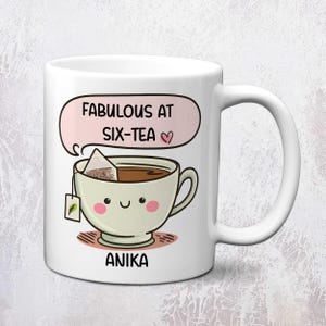 May include: A white ceramic mug featuring a cartoon illustration of a smiling teacup with pink cheeks and a tea bag. Above the cup is a pink speech bubble with the text "FABULOUS AT SIX-TEA" and a small heart. The name "ANIKA" is printed below the cup.