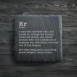 May include: A dark grey slate coaster with the word "Mr" and its definition etched in white. The coaster is placed on a dark wood surface, with the text describing a married man.