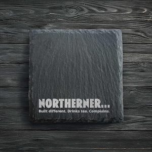 Op de afbeelding: Een vierkante, donkergrijze leistenen onderzetter met het woord "NORTHERNER..." in witte tekst. Daaronder staat de zin "Built different. Drinks tea. Complains." De onderzetter ligt op een donker houten oppervlak.