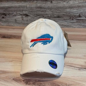 Puede incluir: Una gorra de béisbol blanca con un logotipo de búfalo azul bordado y una franja roja. La gorra tiene una visera desgastada y deshilachada, y una pegatina circular en la parte delantera que dice Vintage. Una pequeña etiqueta de papel está sujeta al lateral.