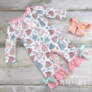 May include: A white long-sleeved baby onesie with a pink and blue print of dresses. The onesie has a pink and white gingham ruffle trim and a bow on each leg. The onesie has a tag that says "Pete + Lucy".