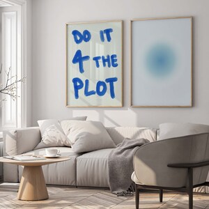 Puede incluir: Un interior de sala de estar con un sofá blanco, una mesa de café de madera y un sillón gris. Dos impresiones enmarcadas cuelgan de la pared. La impresión de la izquierda dice "Do it 4 the plot" en pintura azul. La impresión de la derecha es un diseño abstracto azul y blanco.