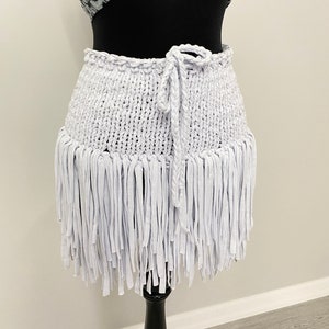 May include: A white, handmade skirt with a knitted waistband and a fringe hem. The skirt is displayed on a black mannequin and features a braided tie closure. The skirt's fringe hangs down, creating a textured, layered effect.