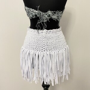 May include: A white, fringed skirt displayed on a black mannequin. The skirt features a woven waistband and a fringe of fabric strips. A patterned scarf is tied around the mannequin's torso above the skirt.