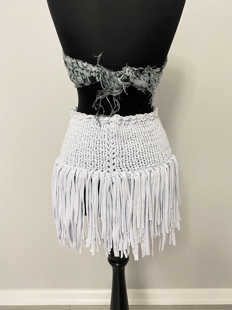 May include: A white, fringed skirt displayed on a black mannequin. The skirt features a woven waistband and a fringe of fabric strips. A patterned scarf is tied around the mannequin's torso above the skirt.