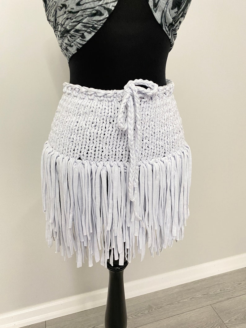 May include: A white, handmade skirt with a knitted waistband and a fringe hem. The skirt is displayed on a black mannequin and features a braided tie closure. The skirt's fringe hangs down, creating a textured, layered effect.