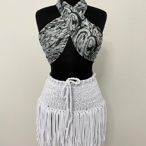 May include: A two-piece outfit on a black mannequin. The top is a halter style with a black and white swirled pattern. The bottom is a white, fringed skirt with a drawstring waist. The outfit is displayed on a black mannequin.