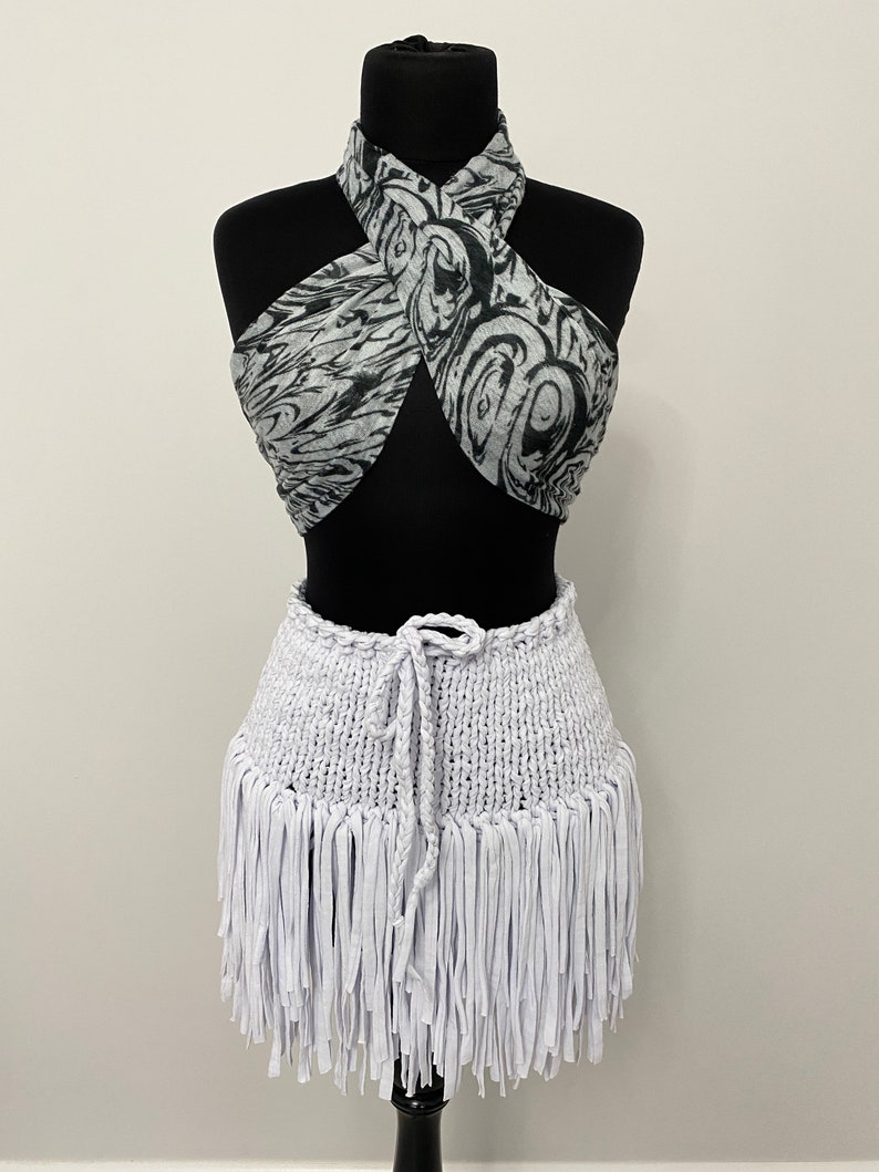 May include: A two-piece outfit on a black mannequin. The top is a halter style with a black and white swirled pattern. The bottom is a white, fringed skirt with a drawstring waist. The outfit is displayed on a black mannequin.