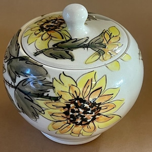 May include: A white ceramic sugar bowl with a lid, decorated with hand-painted yellow sunflowers and green leaves. The bowl is round with a small white knob on the lid. The floral design adds a touch of rustic charm.