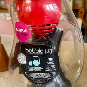 May include: A clear plastic water jug with a red lid and filter. The jug is labelled "bobble jug make water better." The label also says "26 gallons 100 litres" and "includes one filter".