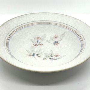 May include: A white ceramic plate with a floral design. The plate has a slightly raised rim with two brown and one blue concentric rings. The floral design features white flowers with green stems and accents of purple and blue.