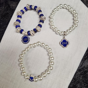 Puede incluir: Tres pulseras de cuentas. Una pulsera presenta cuentas azules y plateadas con un colgante azul. Otra pulsera está hecha de perlas blancas con un dije azul en forma de trébol. La tercera pulsera está hecha de perlas blancas con un dije de pata azul.