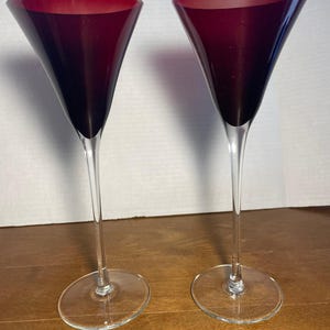 May include: Two elegant wine glasses with a deep burgundy bowl and a clear, slender stem. The glasses have a flared, conical shape and sit on a clear, circular base. The background is a light wood surface.