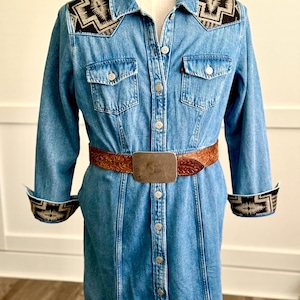 May include: A light blue denim dress with long sleeves, a button-down front, and a brown leather belt with a silver buckle. The dress features decorative black and white patterned accents on the shoulders and cuffs.