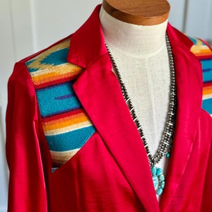 Puede incluir: Una blazer rojo brillante con detalles de hombros estampados coloridos. La blazer se exhibe en un maniquí y se combina con un collar de cuentas plateadas y un colgante turquesa. La blazer tiene solapas clásicas y cierre de botón.