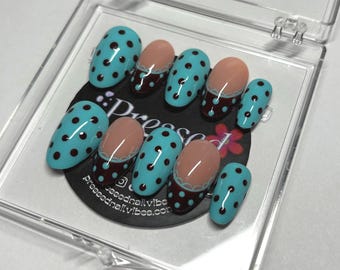 Pode incluir: Um conjunto de dez unhas postiças num estojo de plástico transparente. As unhas apresentam uma base azul-clara com bolinhas castanho-escuras, enquanto algumas têm uma ponta castanho-escura com uma linha festonada branca. O texto Pressed é visível.