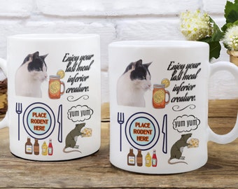 Puede incluir: Dos tazas de cerámica blanca con un diseño de gato blanco y negro. Las tazas muestran un gato mirando un vaso de limonada con una rodaja de limón. El texto en las tazas dice "Enjoy your last meal inferior creature." y "yum yum". Hay un plato con un tenedor y un cuchillo y una rata sosteniendo un trozo de queso en cada taza.