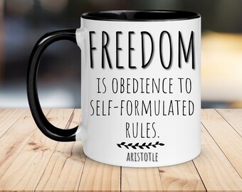 Puede incluir: Taza de cerámica blanca con borde y asa negros. La taza presenta una cita en texto negro: "La libertad es la obediencia a las reglas autoformuladas." La cita se atribuye a Aristóteles.