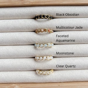 Puede incluir: Cinco anillos de alambre dorado envueltos con piedras preciosas de diferentes colores. Los anillos se muestran sobre un fondo de tela blanca. Las piedras preciosas son obsidiana negra, jade multicolor, aguamarina facetada, piedra lunar y cuarzo transparente.