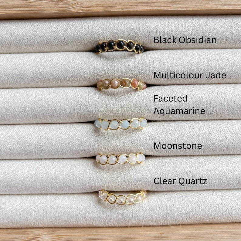 Puede incluir: Cinco anillos de alambre dorado envueltos con piedras preciosas de diferentes colores. Los anillos se muestran sobre un fondo de tela blanca. Las piedras preciosas son obsidiana negra, jade multicolor, aguamarina facetada, piedra lunar y cuarzo transparente.