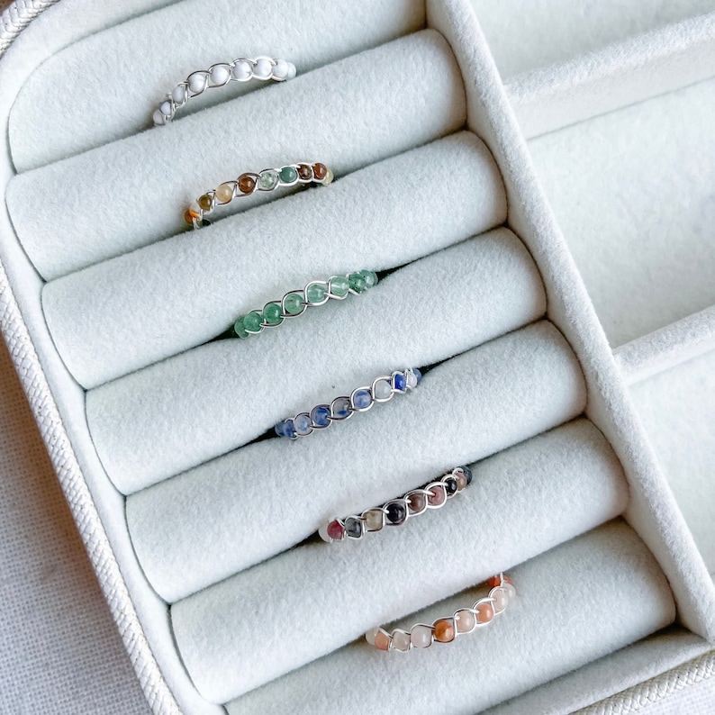 May include: Six rings made of silver wire and various coloured gemstones are displayed in a white velvet ring box. The rings are arranged in a row, with the white gemstone ring at the top and the orange gemstone ring at the bottom.