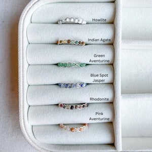 May include: Six gemstone rings displayed in a white ring box. The rings are made with different coloured gemstones, including howlite, Indian agate, green aventurine, blue spot jasper, rhodonite, and pink aventurine.