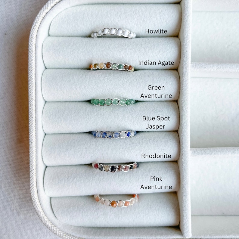 May include: Six gemstone rings displayed in a white ring box. The rings are made with different coloured gemstones, including howlite, Indian agate, green aventurine, blue spot jasper, rhodonite, and pink aventurine.