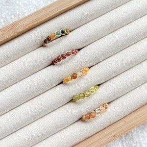 Könnte beinhalten: Fünf golddrahtgewickelte Ringe mit verschiedenen farbigen Edelsteinen. Die Ringe werden in einer Holzkiste mit weißem Samtbezug präsentiert.