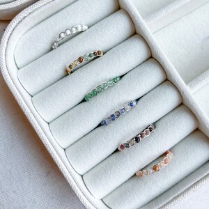 May include: Six rings with different coloured gemstones are displayed in a white ring box. The rings are made of silver wire and feature a variety of gemstones, including white, brown, green, blue, and pink.
