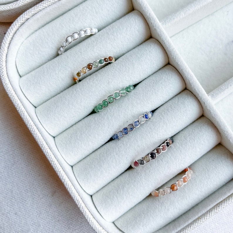 May include: Six rings with different coloured gemstones are displayed in a white ring box. The rings are made of silver wire and feature a variety of gemstones, including white, brown, green, blue, and pink.