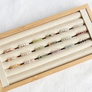 May include: A selection of gemstone rings presented in a wooden tray. The rings showcase a variety of coloured stones, such as pink, green, and purple, set in a silver wire design. The rings are arranged in rows on a beige surface.