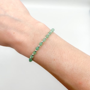 May include: A bracelet with small, round, light green beads and silver-coloured beads. The bracelet is strung on a thin cord and is designed to be worn on the wrist. The beads are evenly spaced. The bracelet is a piece of jewellery.