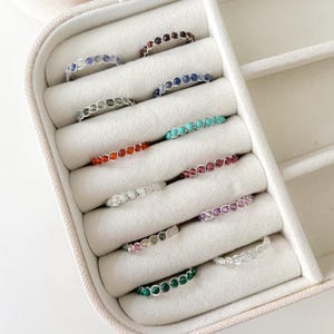 May include: A collection of 10 gemstone rings displayed in a ring box. The rings are made with different coloured gemstones, including red, orange, yellow, green, blue, purple, and white.