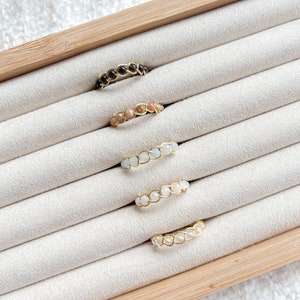Puede incluir: Cinco anillos de alambre dorado con piedras preciosas de diferentes colores. Los anillos se muestran sobre un fondo de terciopelo blanco en una caja de madera.