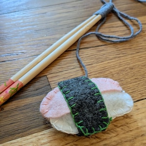 May include: A felt sushi ornament with pink, white, and gray components, stitched with green thread. The ornament is attached to a pair of wooden chopsticks with floral designs and a gray string.
