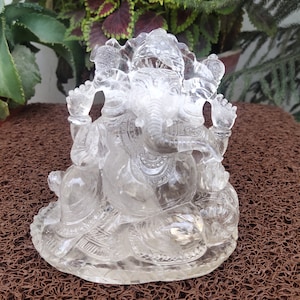 May include: A clear crystal sculpture of the Hindu deity Ganesha, sitting with his trunk raised and holding a sweet in his hand. The sculpture is intricately carved with details of Ganesha's traditional attire and jewelry.