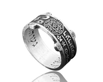 May include: Silver ring with engraved symbols and text. The ring features a central circular design with a star, surrounded by other symbols and inscriptions. The band is wide and has a detailed, textured surface.
