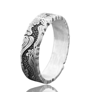May include: A silver ring with an ornate, flowing pattern. The design features swirling lines and small, textured details, creating a detailed and artistic look. The ring's band has a textured edge. The ring is displayed against a white background.