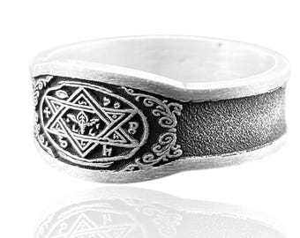 May include: A silver-toned ring with an engraved design. The ring features a central oval with a star and other symbols, surrounded by ornate scrollwork. The band has a textured, dark gray surface.