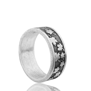 May include: A silver ring featuring a repeating maple leaf pattern. The wide band displays a detailed design of leaves and textured lines. The ring is presented against a white backdrop.