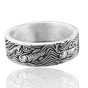 May include: A silver ring with a detailed aquatic design. The ring features a repeating pattern of fish swimming through stylized waves. The band is wide and has a slightly rounded shape, with the design wrapping around the entire circumference.