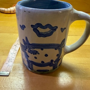 May include: A white ceramic mug with a blue interior and a blue hand-painted design featuring a cow and a cloud-like shape. A measuring tape placed next to the mug indicates a height of approximately 3.5 inches for the main body of the mug.
