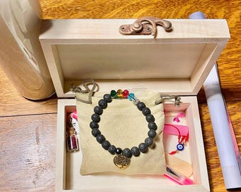 May include: A wooden box with a hinged lid, containing a black lava rock bracelet with a gold tree of life charm, a small glass bottle with a cork stopper, a pink and white fabric pouch, a red string with a blue evil eye charm, and a pink and white fabric strip.